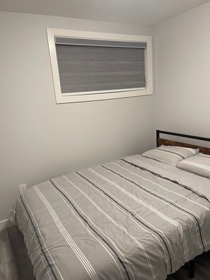 A bedroom with a bed, a window with blinds, and a wall-mounted light switch. - Brighton basement suite with 1 bedroom + sleeper sofa, peaceful and private. (Saskatoon)