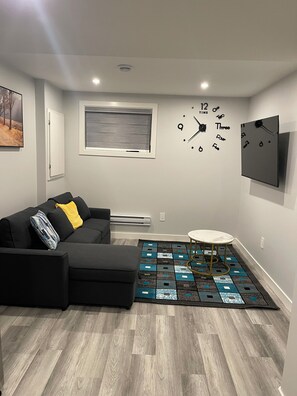 A modern living room with a sectional sofa, a small round table, a clock, and a flat-screen TV mounted on the wall. - Brighton basement suite with 1 bedroom + sleeper sofa, peaceful and private. (Saskatoon)
