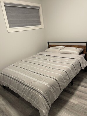 A single bed with a wooden headboard, a grey striped bedspread, and a window with blinds. - Brighton basement suite with 1 bedroom + sleeper sofa, peaceful and private. (Saskatoon)