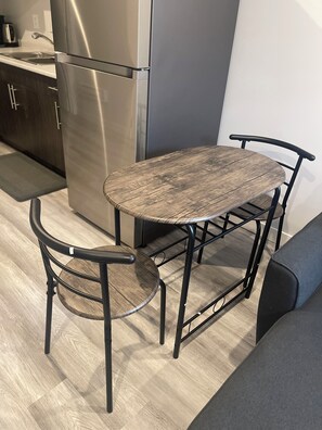 A kitchenette with a stainless steel refrigerator, a wooden dining table with metal chairs, and a grey sofa. - Brighton basement suite with 1 bedroom + sleeper sofa, peaceful and private. (Saskatoon)