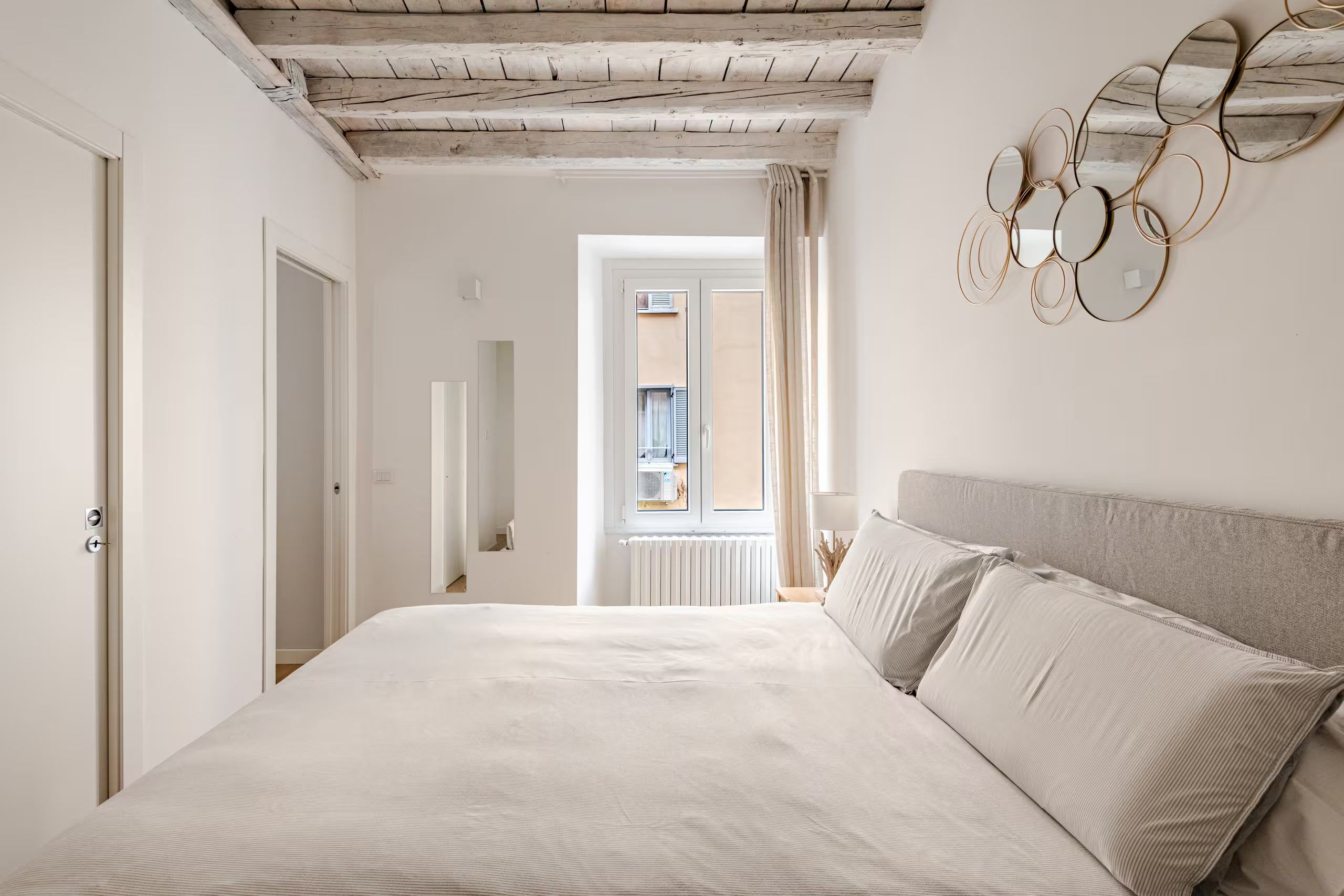 A bedroom with a large bed, a wooden ceiling, a view through a window, and a wall-mounted mirror.