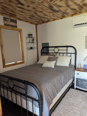 Ein Schlafzimmer mit einem Bett, einem Nachttisch, einem Wandregal, einem Fenster mit Jalousien und einer Holzbalkendecke.