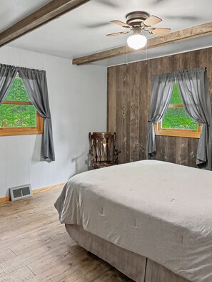 A bedroom with a large bed, wooden beams, a ceiling fan, and two windows with curtains. - Cozy 2-bedroom cottage in serene Lakewood (Lakewood)