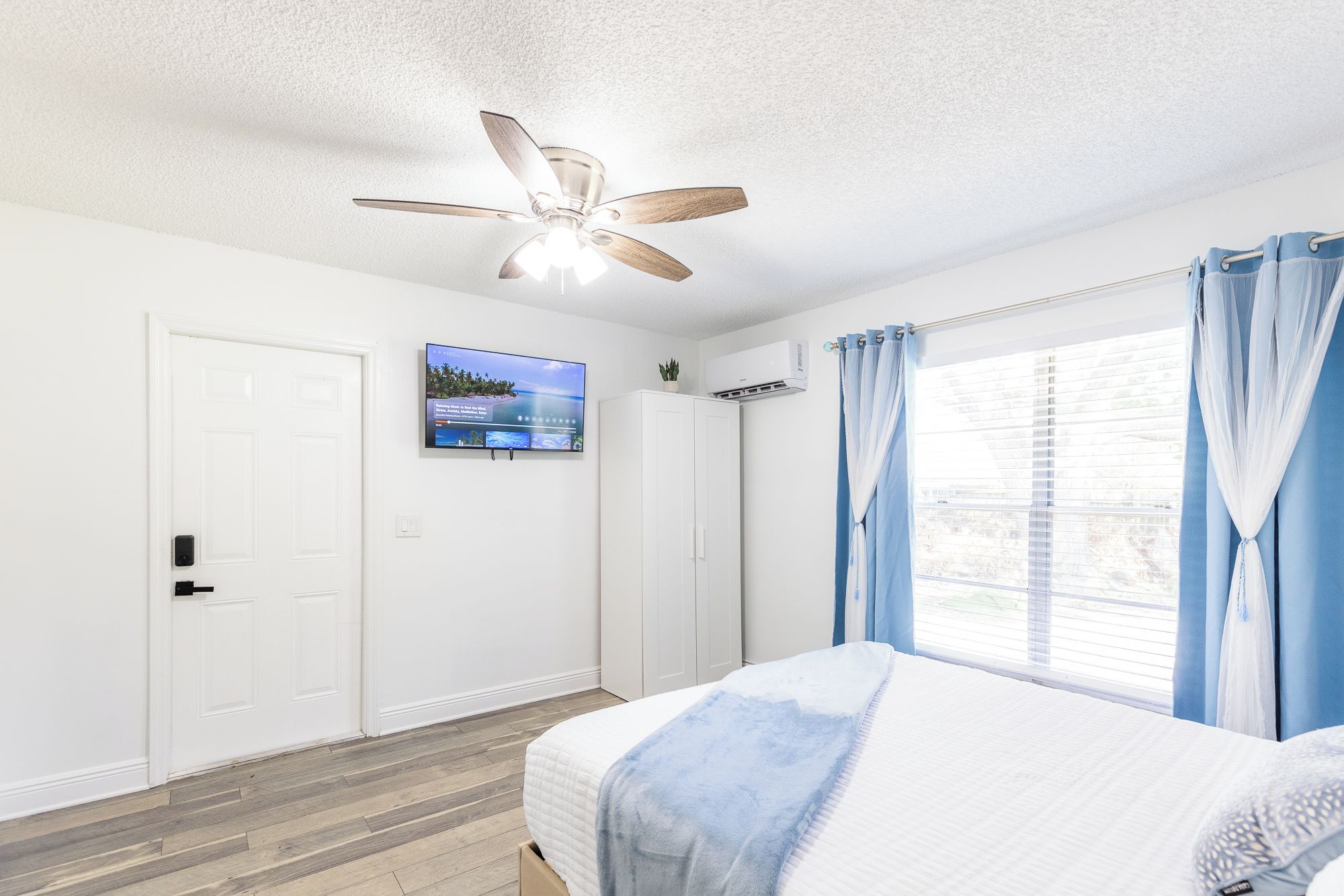 A bedroom with a bed, a ceiling fan, a TV on the wall, and a window with curtains.