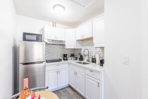 Una cocina compacta con armarios blancos, un refrigerador de acero inoxidable, un microondas y una cafetera.