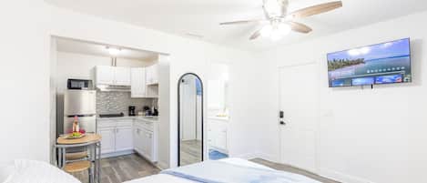 A compact hotel room with a bed, a ceiling fan, a TV, and a kitchenette.