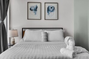 A hotel room with a bed, two framed abstract paintings, a bedside lamp, and a rolled towel on the bed. - Luxury 1BR + Den | Bay & Pool Views | Brickell (Miami)