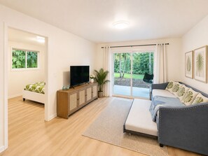 A living room with a sofa, a television on a wooden cabinet, a large sliding glass door to a garden, and a bed with a green leaf-patterned bedspread. - Palm Paradise — AC + Gym + Game Room Sleeps 15 (Keaau)