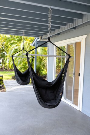 A black hammock suspended from ceiling beams in a covered outdoor area with a view of a house and palm trees. - Palm Paradise — AC + Gym + Game Room Sleeps 15 (Keaau)