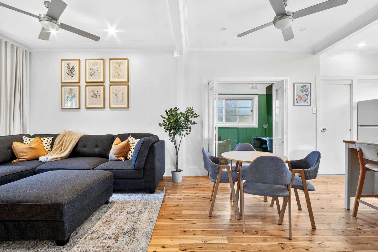 A modern living room with a sectional sofa, dining table, and chairs, a potted plant, and framed botanical prints on the wall.
