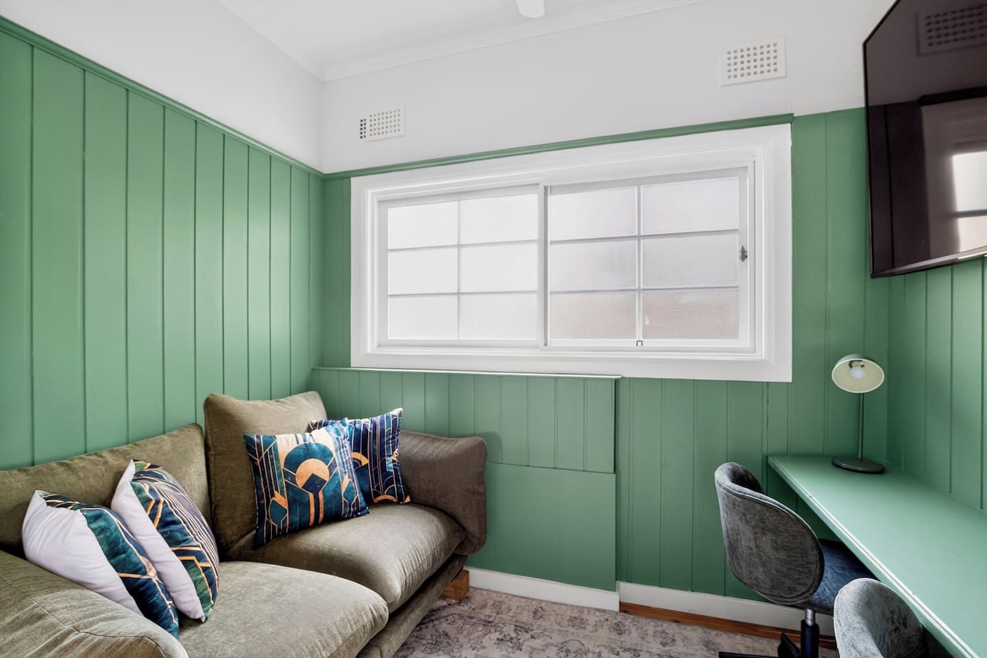 A room with a green wall, a window, a sofa with cushions, a desk, and a chair.