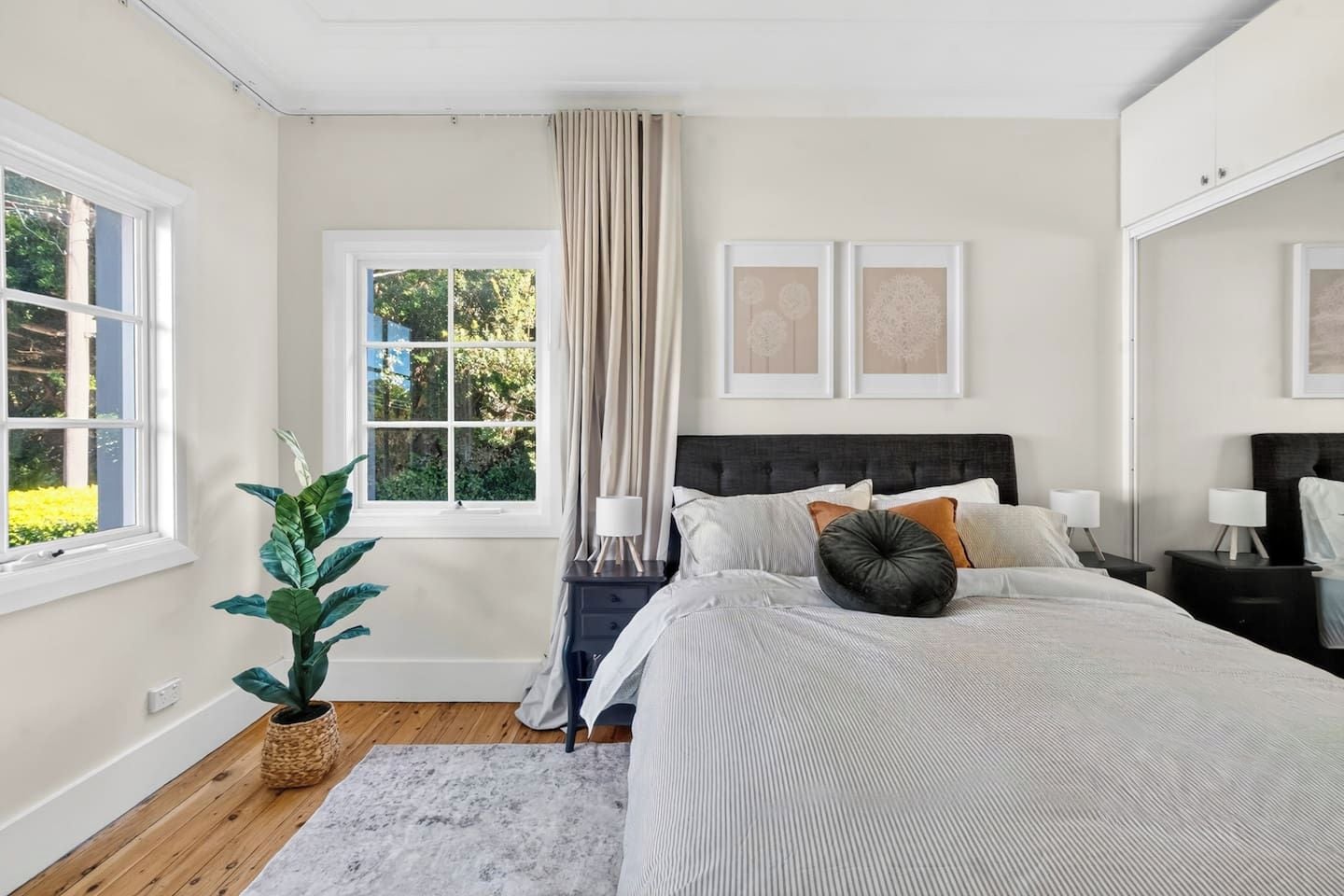 A bedroom with a large bed, a window with curtains, a bedside table with a lamp, and a potted plant.