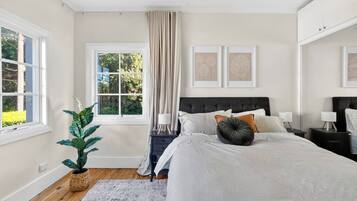 A bedroom with a large bed, a window with curtains, a bedside table with a lamp, and a potted plant.