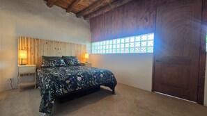 Un dormitorio con una cama, dos mesitas de noche con lámparas, un cabecero de madera, un ventanal con vidrio decorativo y una puerta de madera.