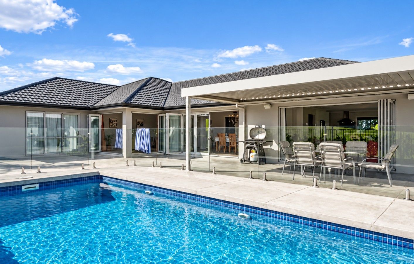 Una piscina con solárium empedrado, mobiliario exterior y un patio cubierto.
