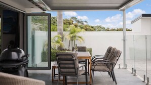 Un patio exterior con una mesa y sillas, un vidrio que da a un jardín, y una vista despejada del cielo y la vegetación.