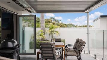 Un patio exterior con una mesa y sillas, un vidrio que da a un jardín, y una vista despejada del cielo y la vegetación.