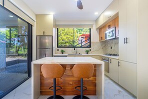 Cozinha moderna com uma ilha grande, dois banquetas, geladeira, fogão e janela com vista para o verde.