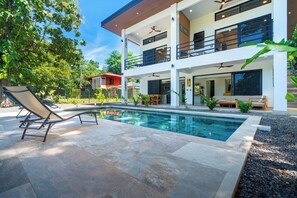 Ein modernes, zweistöckiges Haus mit Swimmingpool, Sitzgelegenheiten im Freien und einem Balkon.