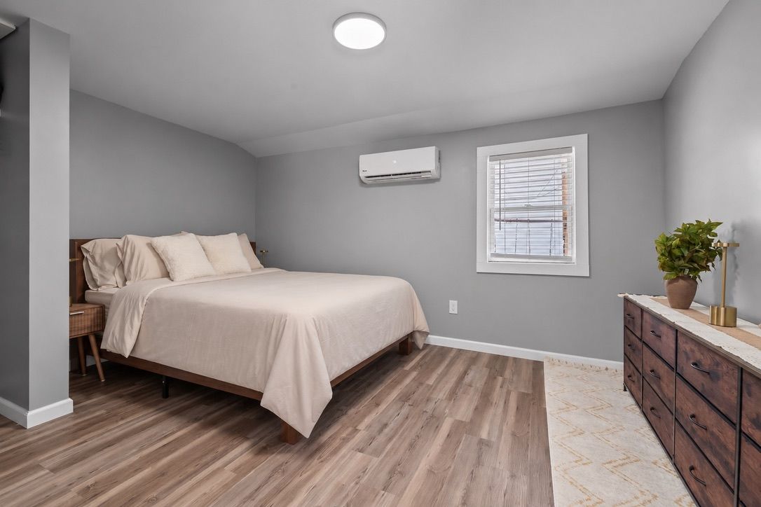 A bedroom with a bed, a dresser, an air conditioning unit, and a window with blinds.