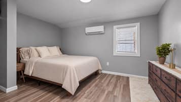 A bedroom with a bed, a dresser, an air conditioning unit, and a window with blinds.