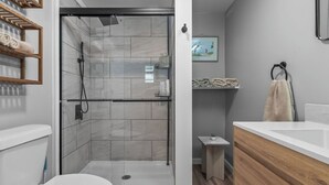 A modern bathroom with a glass-enclosed shower, a white toilet, a wooden vanity, and a small shelf with towels. - The Howder House / Refined 1BR  / Sleeps 4 (Hillsdale)