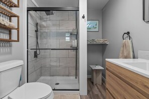 Une salle de bain moderne avec une douche à parois de verre, des toilettes blanches, un meuble-lavabo en bois et une petite étagère avec des serviettes.