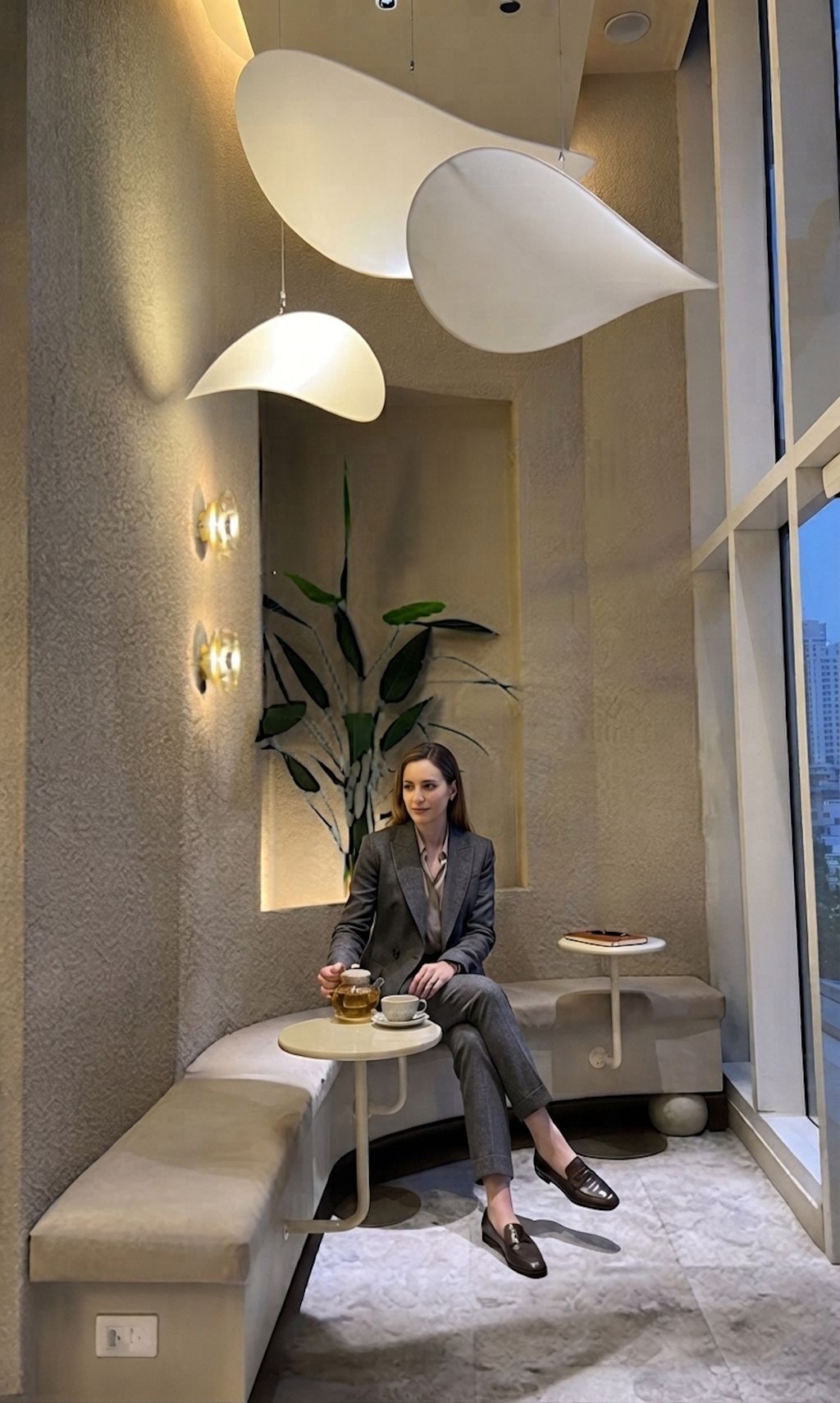 Une personne assise sur un banc dans un intérieur moderne, avec une plante, une table sur laquelle se trouve une tasse et une fenêtre donnant sur un paysage.