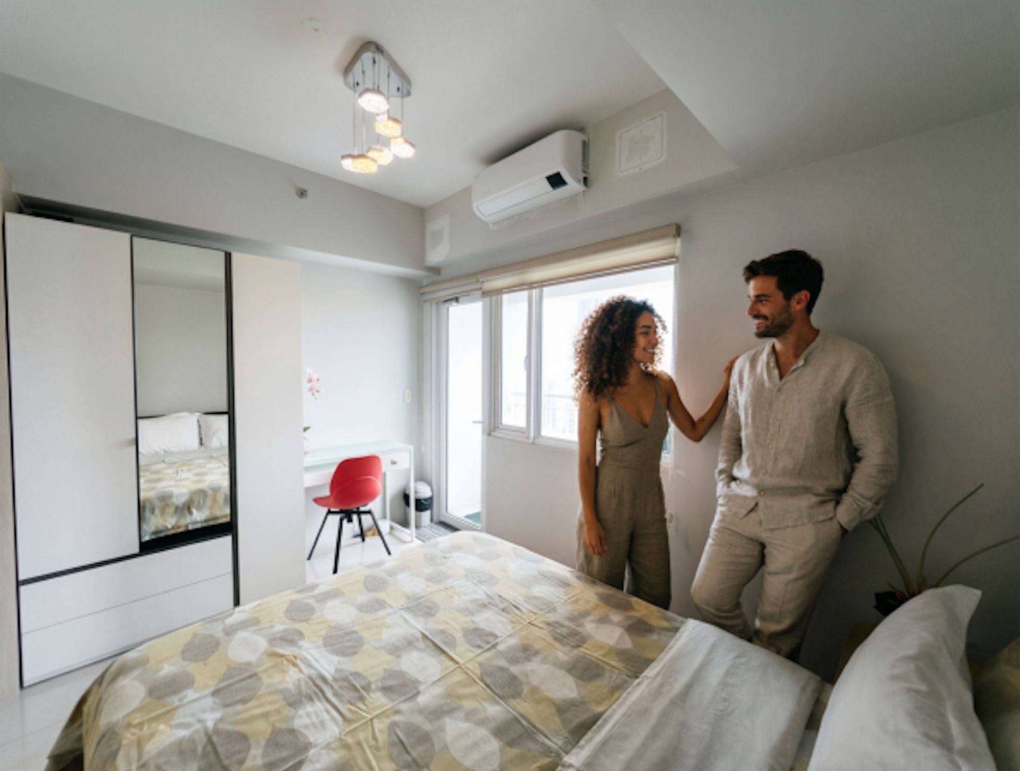 Un dormitorio con una cama, una silla roja, un espejo grande y un equipo de aire acondicionado.