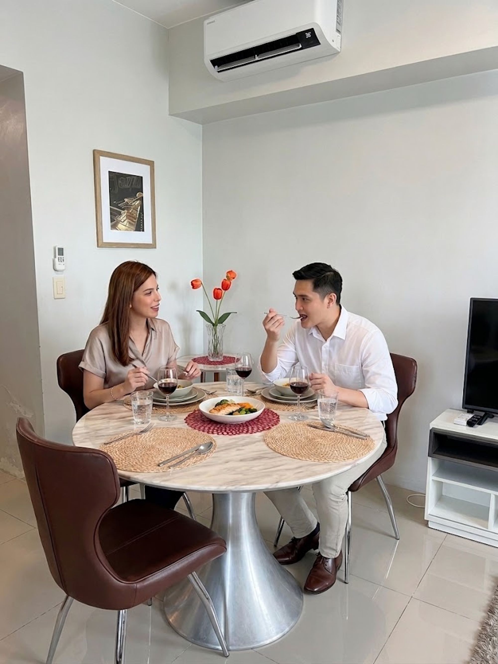 Una mesa de comedor con cubierta de mármol blanco, rodeada de sillas marrones. Hay un jarrón con flores rojas sobre la mesa, y un cuadro en la pared.