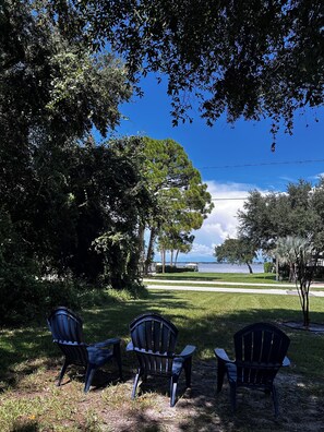 A view of a grassy area with three Adirondack chairs facing a body of water, surrounded by trees and a clear sky.