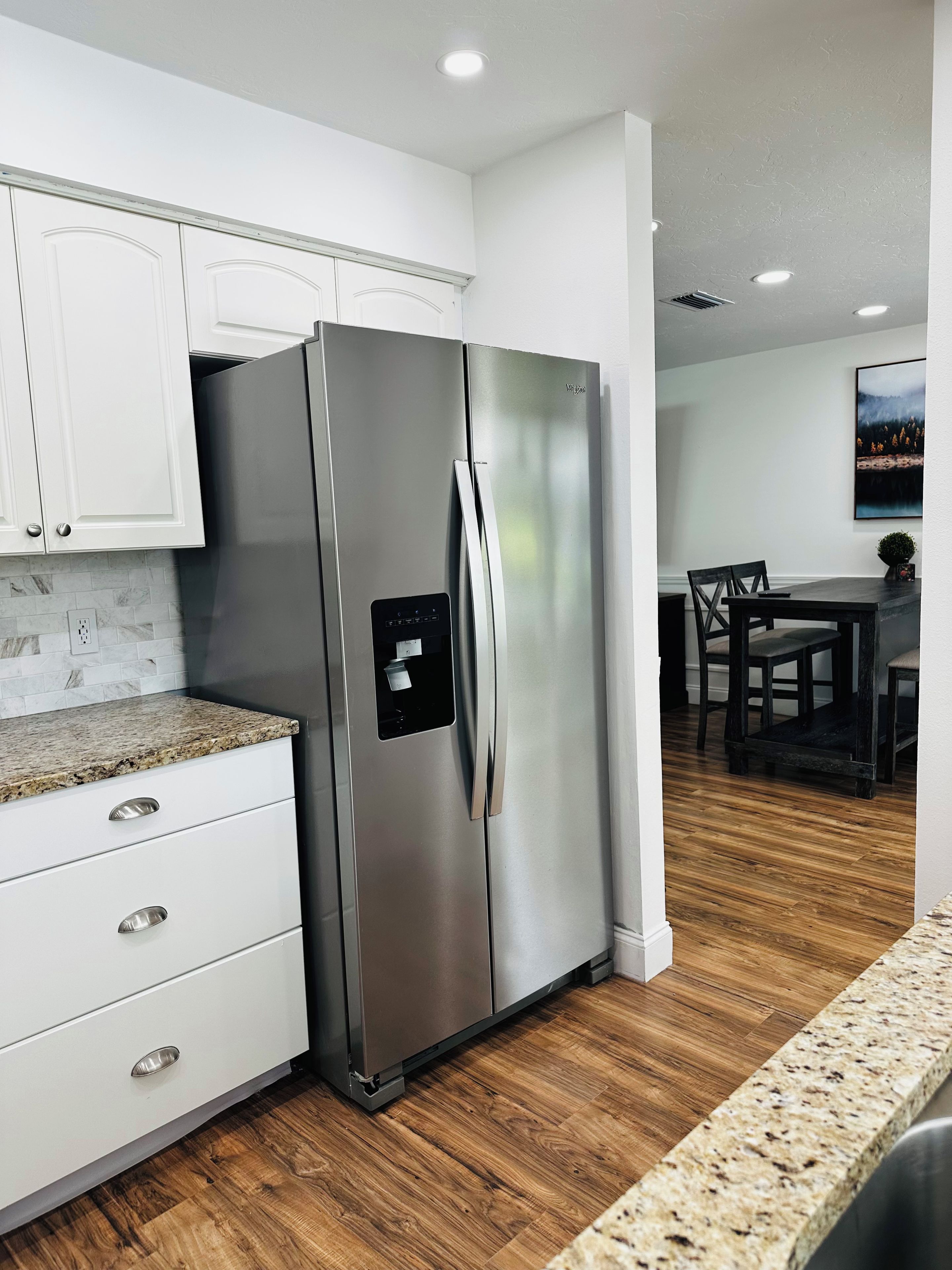 A modern kitchen with a stainless steel refrigerator, white cabinetry, granite countertop, and a dining area with a wooden table and chairs.