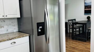 A modern kitchen with a stainless steel refrigerator, white cabinetry, granite countertop, and a dining area with a wooden table and chairs.