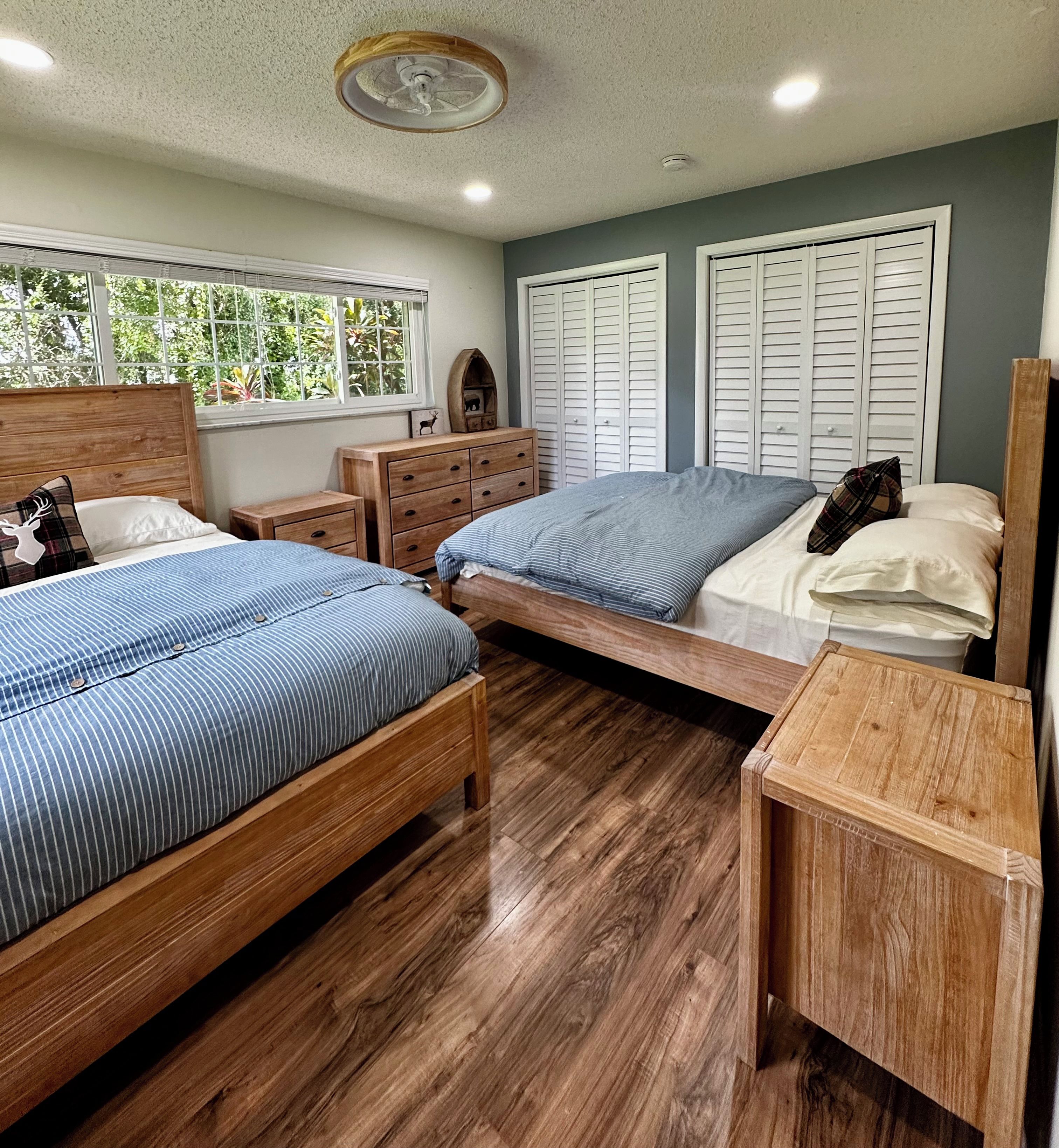 Quarto com duas camas de madeira, uma cômoda e uma janela com vista para o verde.