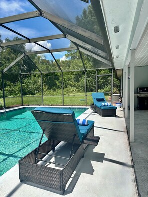 A covered outdoor swimming pool area with lounge chairs and a barbecue grill.