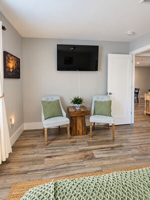 Ein Zimmer mit einem an der Wand befestigten Flachbildfernseher, zwei helle Stühle mit grünen Kissen, einem kleinen Holztisch und einem gewebten Teppich auf dem Boden.