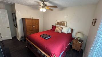 A bedroom with a bed, a wooden wardrobe, a ceiling fan, a nightstand with a lamp, and a window with blinds.