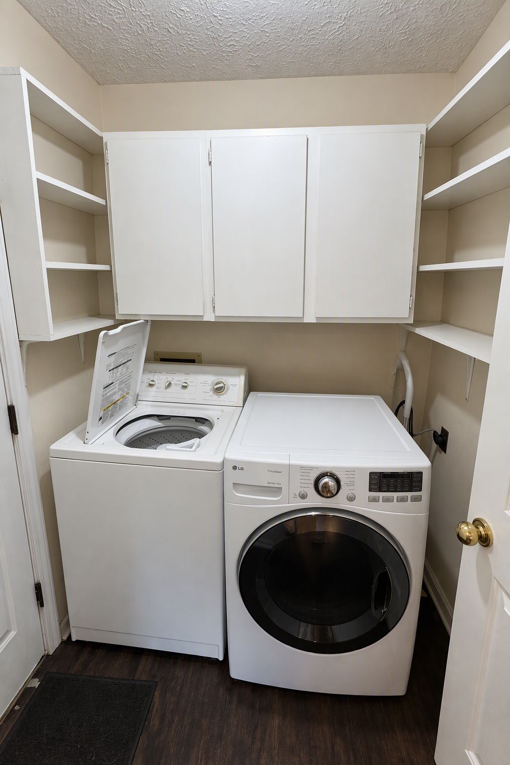 Een wasruimte met een voorlader wasmachine en droger, witte kasten erboven en een zwarte deurmat.