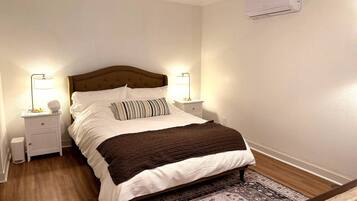A bedroom with a bed, bedside tables, lamps, a rug, and an air conditioning unit.