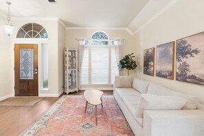 Living room with graceful arched windows, soft neutral tones, a generous white sectional, patterned area rug, sculptural coffee table, and elegant landscape artwork, creating a warm and inviting space. - 21 mins ACL! Executive 4BR Home w/ Office & Yard (Austin)