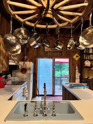 A kitchen with a wooden ceiling, hanging pots, a sink with three faucets, and a view through a glass door. - 1895 Cottage At Belle Knowle (Eldred)