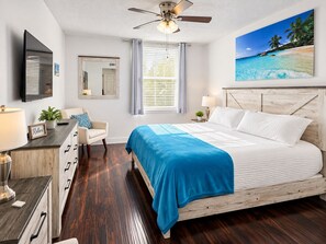 Een slaapkamer met een bed, een bureau, een stoel, een plafondventilator, een raam met zonwering en een schilderij van een strandgezicht.