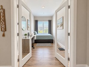 Ein Schlafzimmer mit einem Bett, einem Schreibtisch und einem Stuhl. Es gibt ein Fenster mit Vorhängen, einen Wandspiegel und ein Willkommensschild.