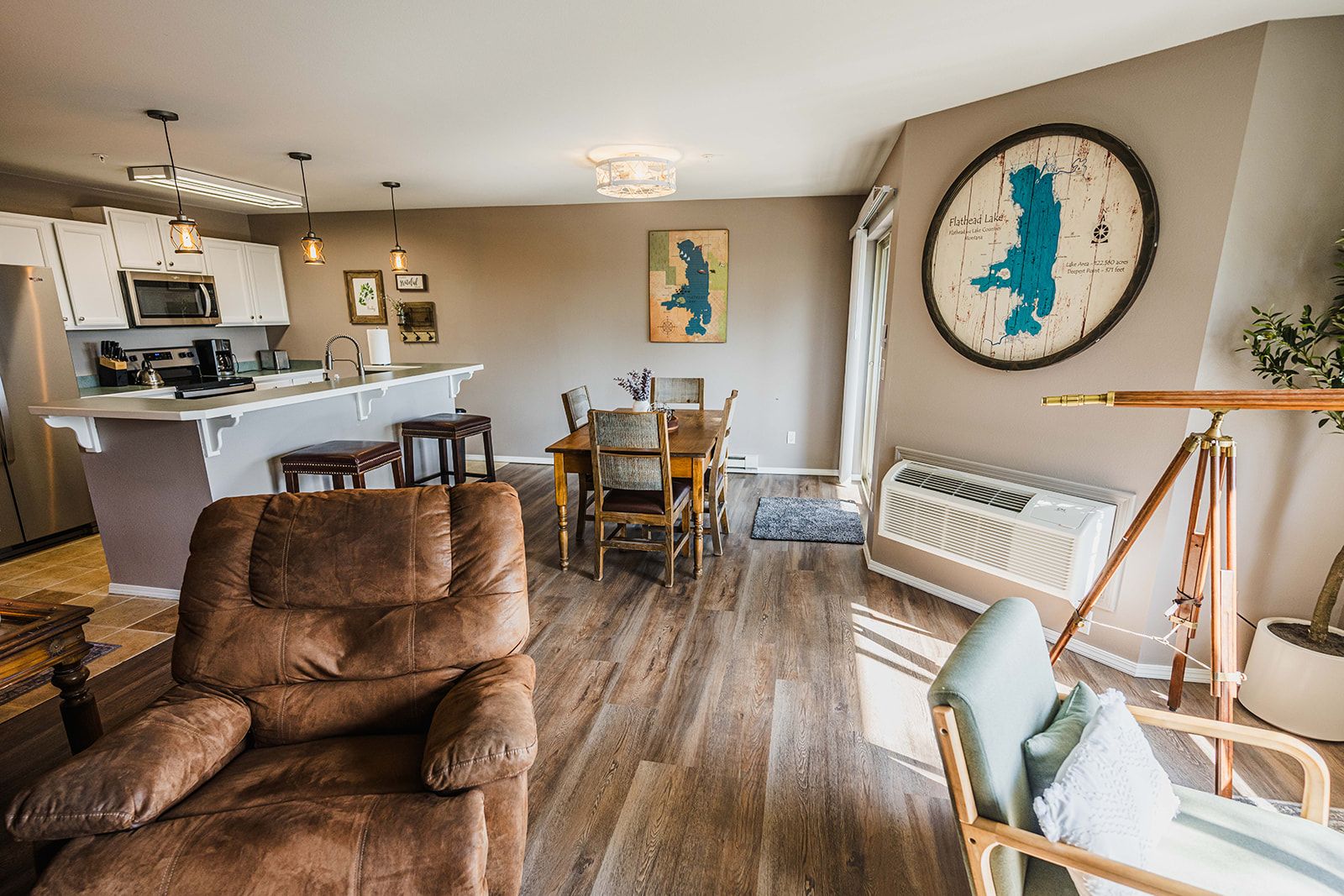 A living room with a brown leather sofa, a dining area with a wooden table and chairs, a kitchen with white cabinets and a microwave, and a large map on the wall.