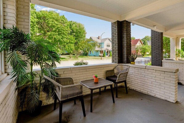 A covered porch with two wicker chairs and a small table, a potted plant, and a brick chimney.