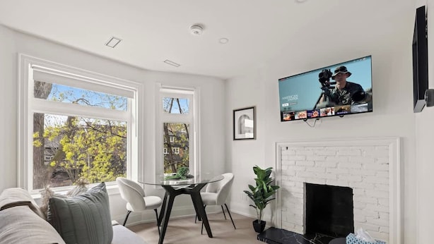 Una sala de estar con chimenea, un televisor montado en la pared y un comedor con mesa de vidrio y sillas.