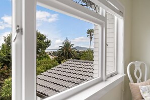 A room with a window overlooking a tiled roof, a white chair, and a view of a landscape with trees and a distant mountain. - The Rose Cottage by Homello (Auckland)
