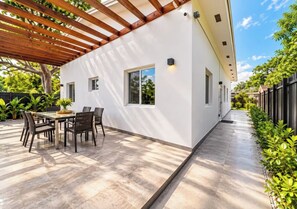 A modern patio with a dining table and chairs, a pergola, and a white exterior wall with windows.