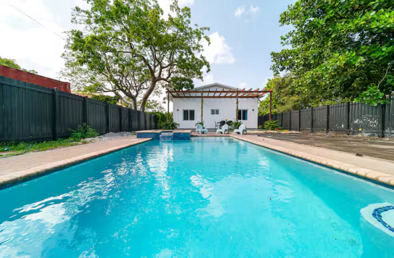 Piscina com uma casa branca e um pergolado ao fundo.