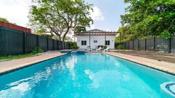 Piscina com uma casa branca e um pergolado ao fundo.